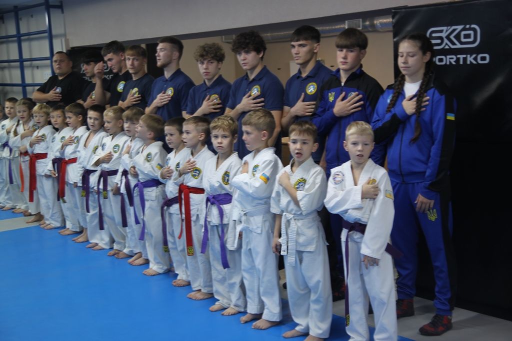 Групове фото юних спортсменів у кімоно та тренерів у спортивній залі. Усі стоять із рукою на грудях.