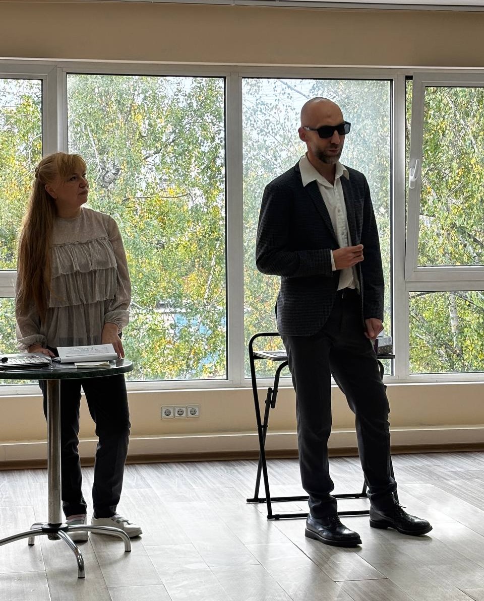 Чоловік у темних окулярах і костюмі виступає біля великого вікна. Поруч стоїть жінка біля столика. За вікном видно зелені дерева.