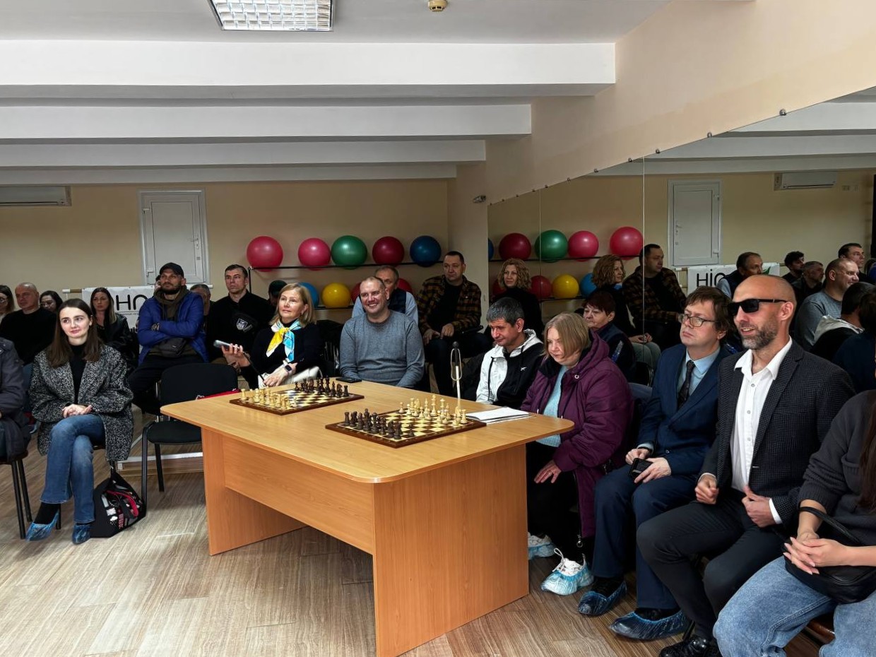 Група людей сидить у залі навколо дерев'яного столу з двома шахівницями. На задньому плані видно дзеркало та різнокольорові фітболи.