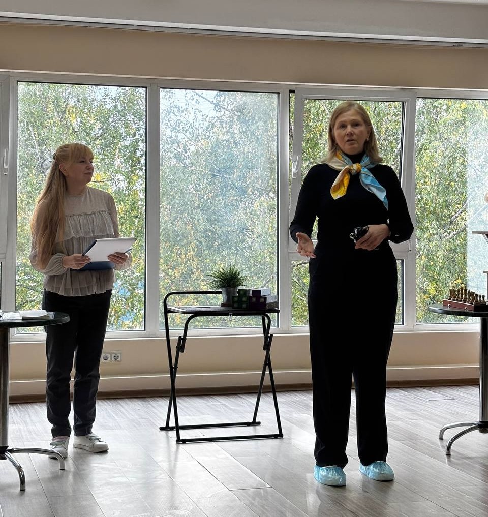 Жінка в чорному костюмі та жовто-блакитній хустці виступає біля панорамного вікна. Поруч стоїть жінка з блокнотом. За вікном — зелені дерева.