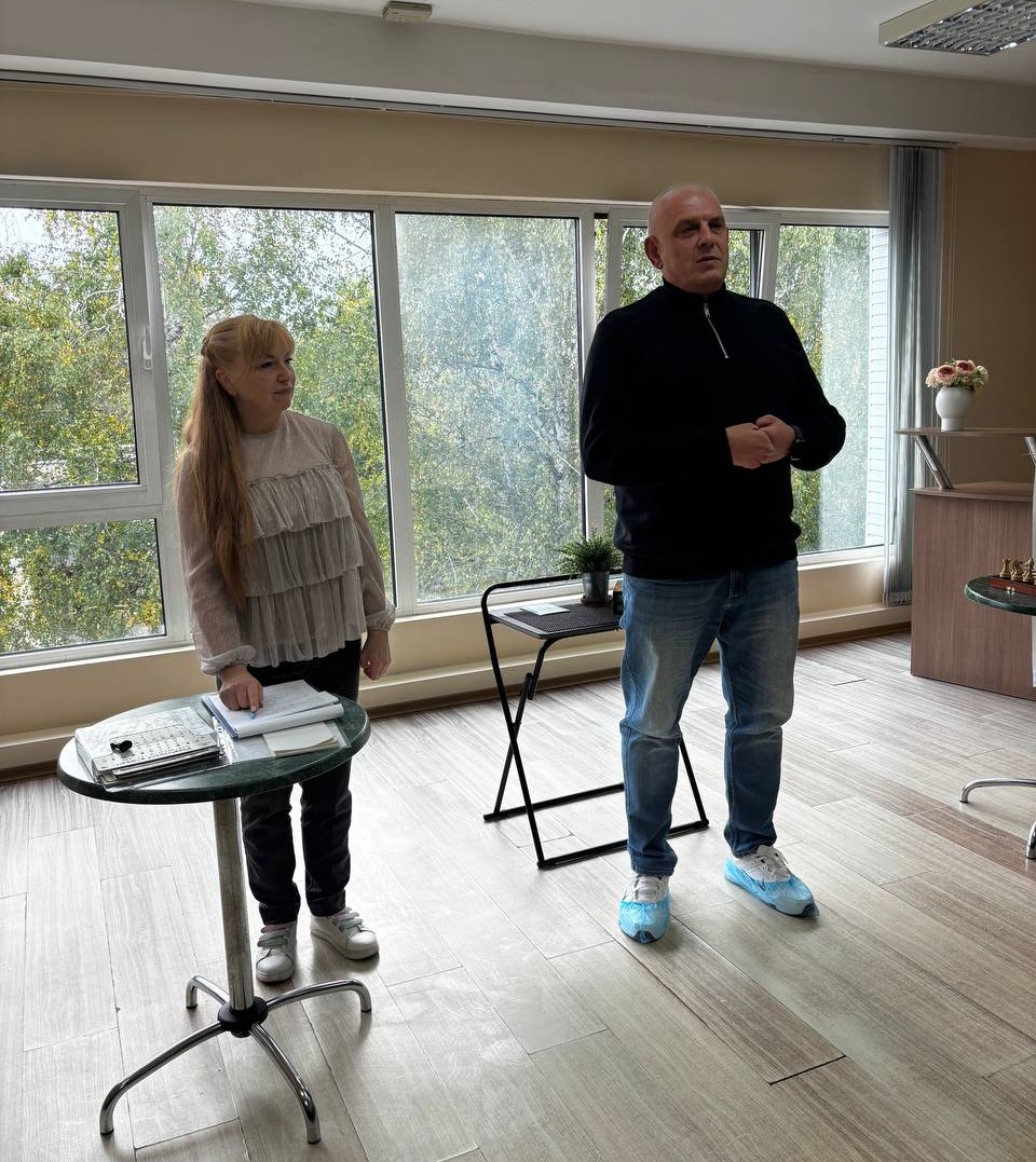 Лисий чоловік у чорній кофті та джинсах виступає біля великого вікна. Поруч стоїть жінка з блокнотом біля круглого столика. За вікном — дерева.