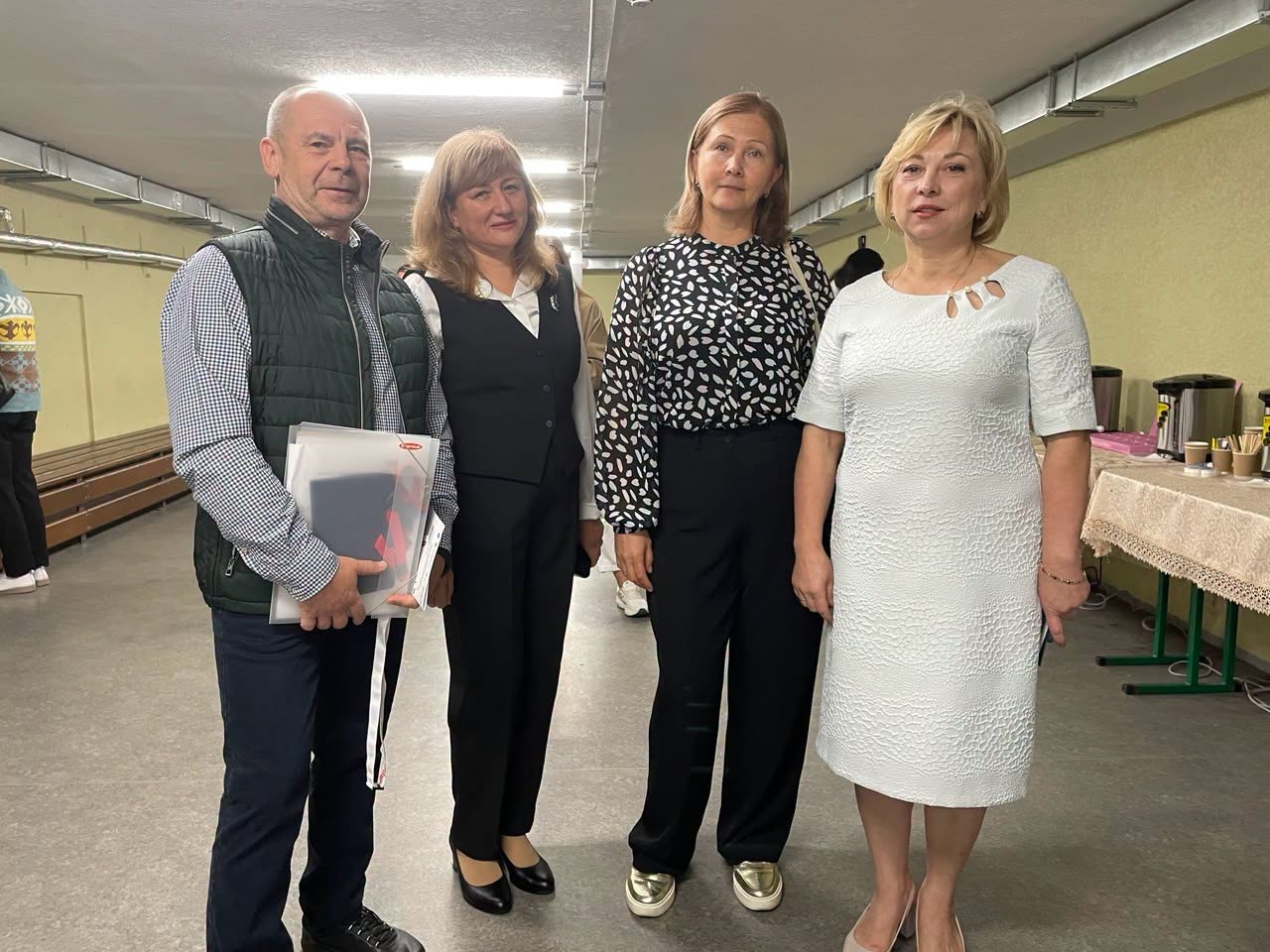 Чотири людини стоять у коридорі та дивляться в кадр. Праворуч видно столик із кавою. На задньому плані вздовж стіни — дерев'яні лави.