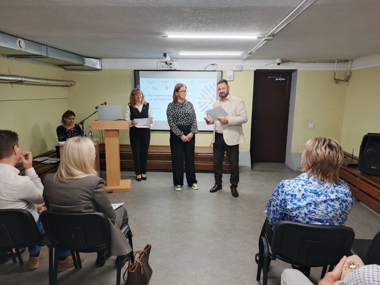 Чоловік у світлій сорочці виступає біля інтерактивної дошки перед слухачами. Люди сидять у два ряди на чорних стільцях.
