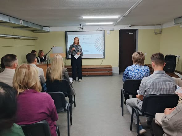 Жінка виступає перед аудиторією в залі з низькою стелею. Позаду неї — інтерактивна дошка. Слухачі сидять на стільцях обличчям до неї.