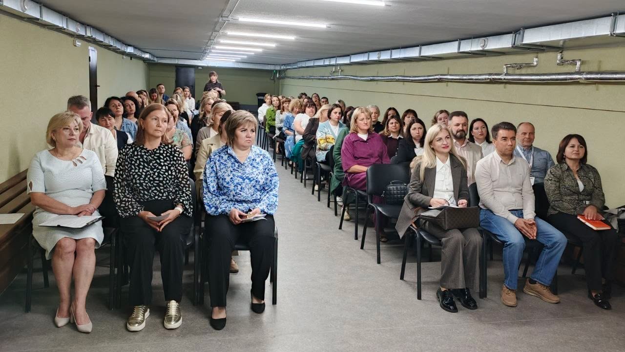 Численна аудиторія сидить у залі з низькою стелею. Стільці розставлені у два великі блоки, між якими є прохід по центру.