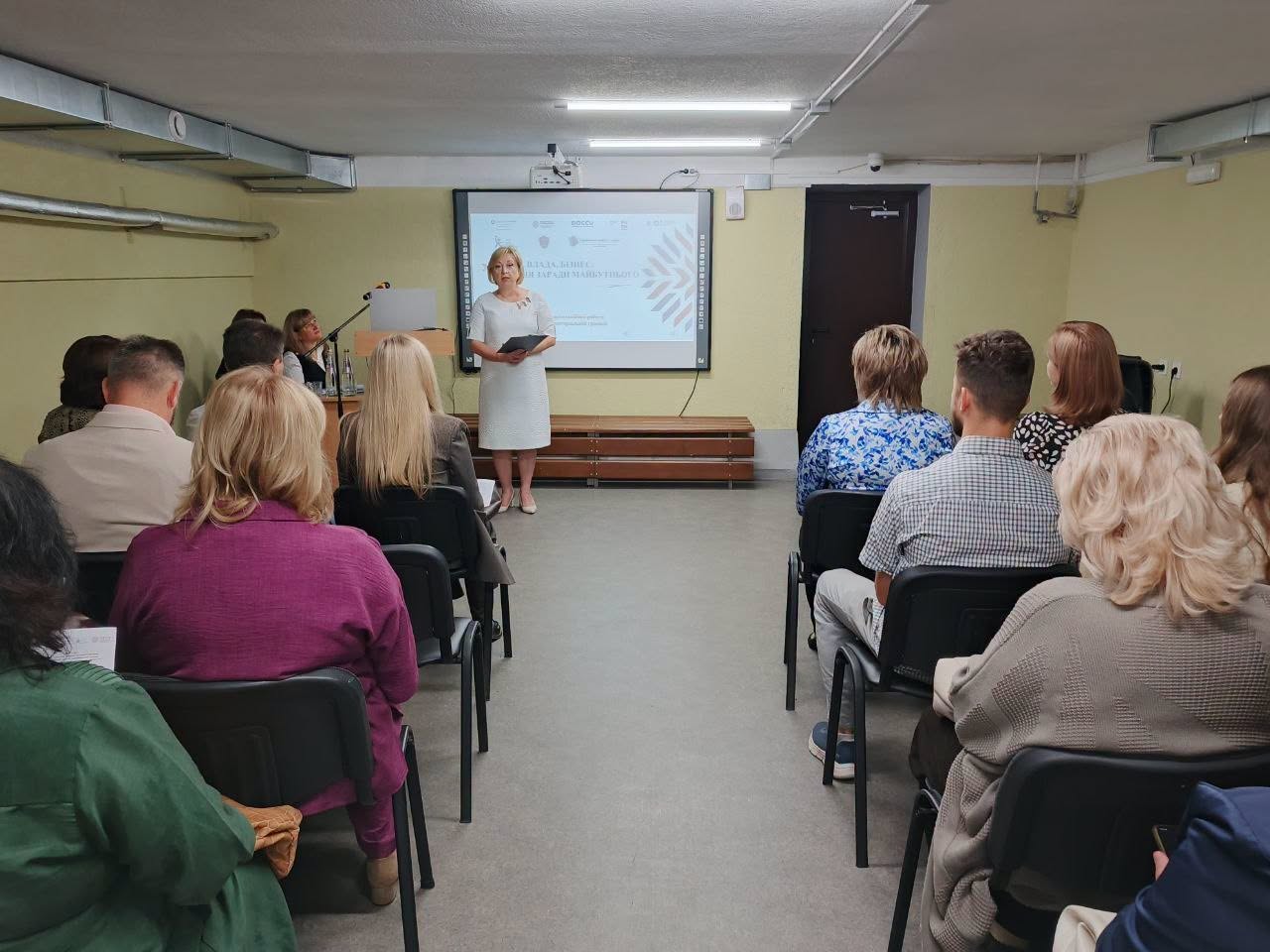 Жінка у білій сукні виступає перед аудиторією у залі. Позаду неї — екран із презентацією. Слухачі сидять у два ряди на стільцях.