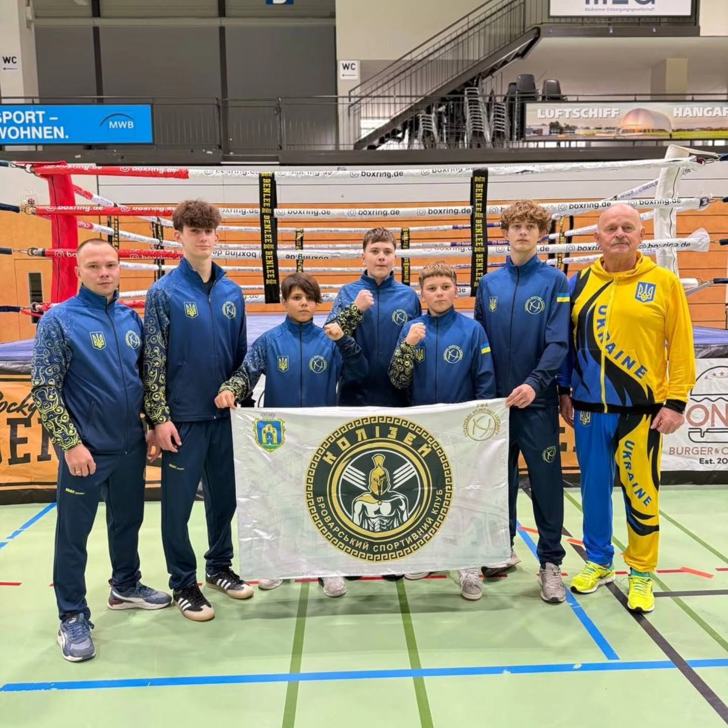 Група спортсменів у синій формі України та тренер у жовтому костюмі позують біля рингу. Вони тримають білий прапор спортивного клубу «Колізей».