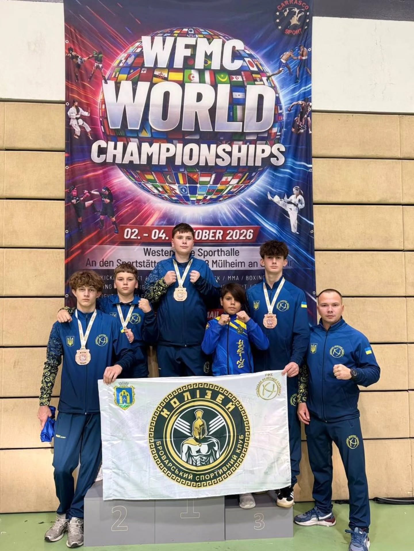 Спортсмени в синій формі з медалями стоять на п'єдесталі під банером чемпіонату світу. Вони тримають білий прапор клубу «Колізей».