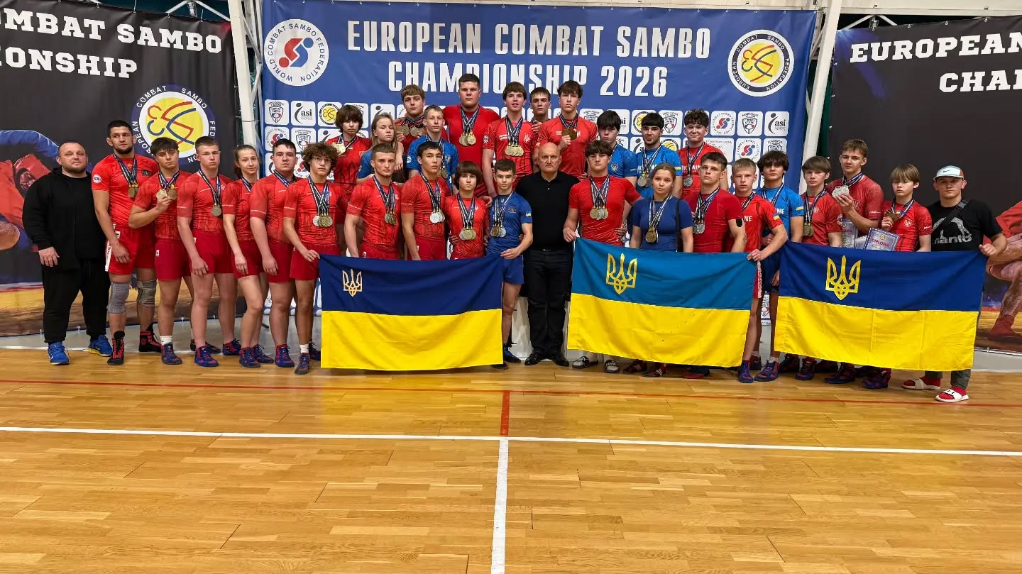Збірна України з бойового самбо: велика група молодих спортсменів із медалями тримає три синьо-жовті прапори на тлі банера чемпіонату Європи.