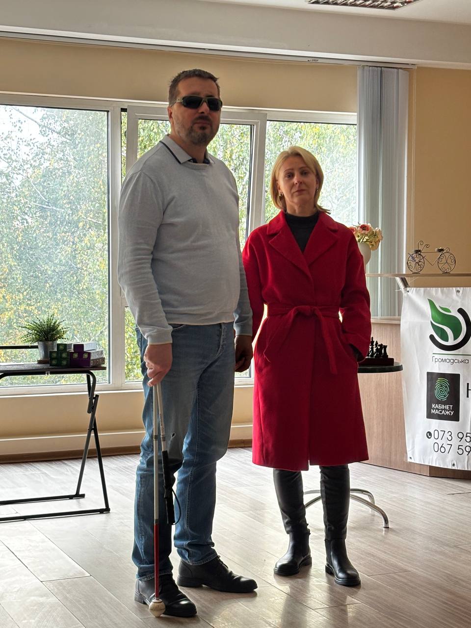 Чоловік у темних окулярах із білою тростиною та жінка в червоному пальті стоять разом у залі. Позаду видно панорамне вікно та банер.