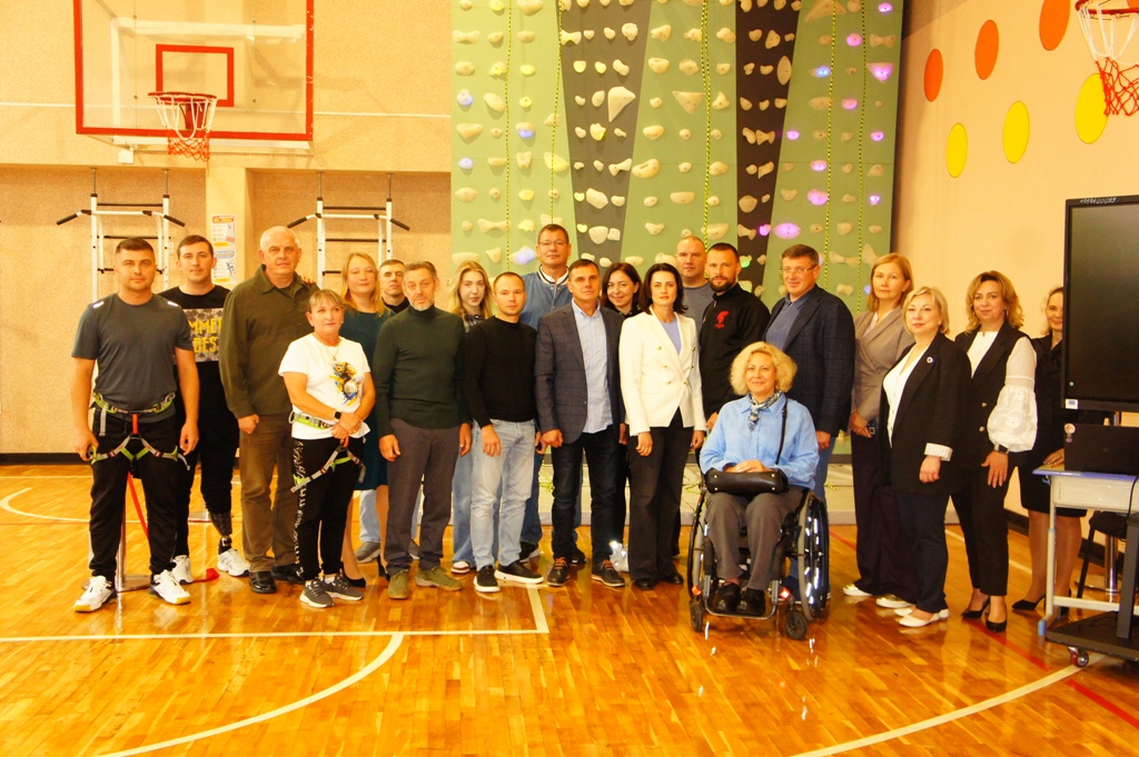 Група людей позує у спортзалі на тлі скеледрому та баскетбольного кільця. Попереду сидить жінка у колісному кріслі.