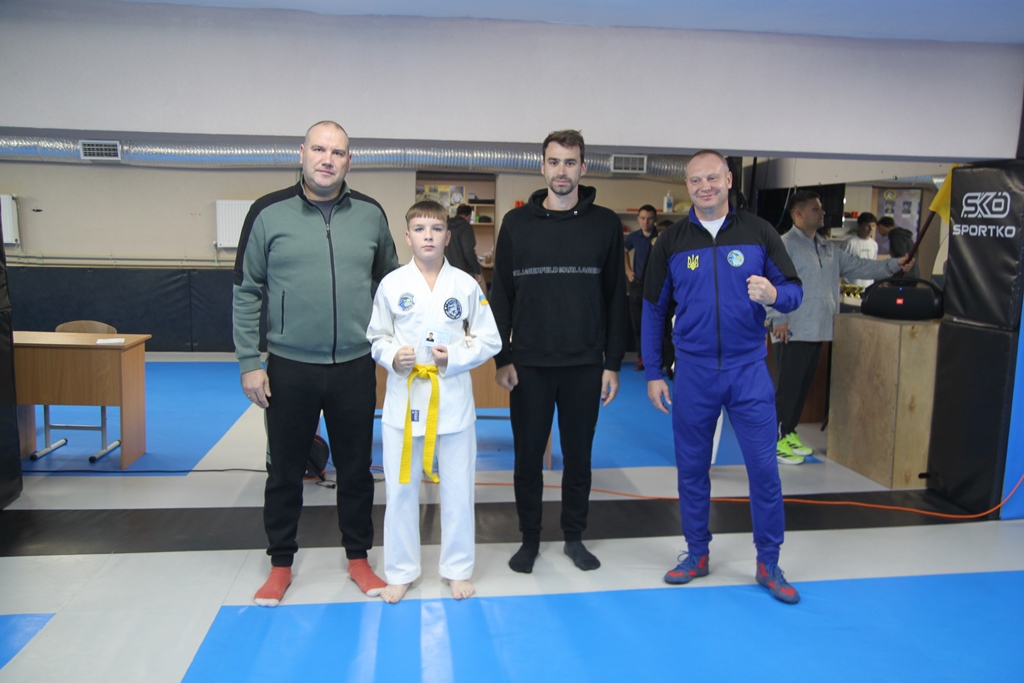 Групове фото у спортзалі. Хлопчик у білому кімоно з жовтим поясом стоїть по центру між трьома дорослими чоловіками. На підлозі синє покриття.