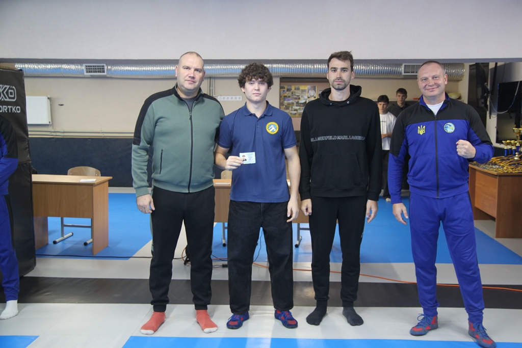 Четверо чоловіків позують у спортивній залі; один тримає посвідчення, інший підняв великий палець.
