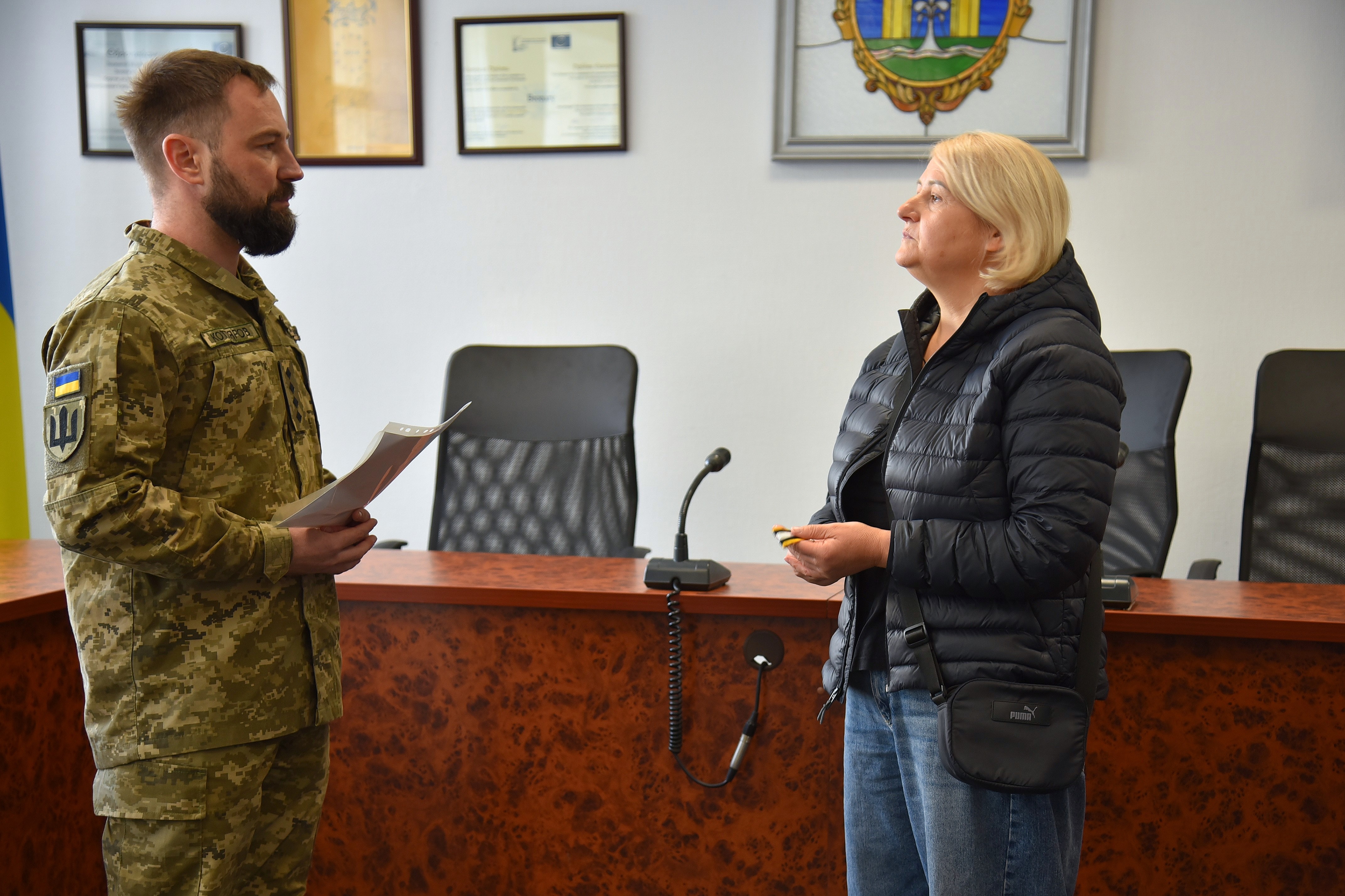 Зблизька: військовий із документами в руках стоїть профілем і спілкується з жінкою у темній куртці. На стіні позаду видно герб і грамоти.