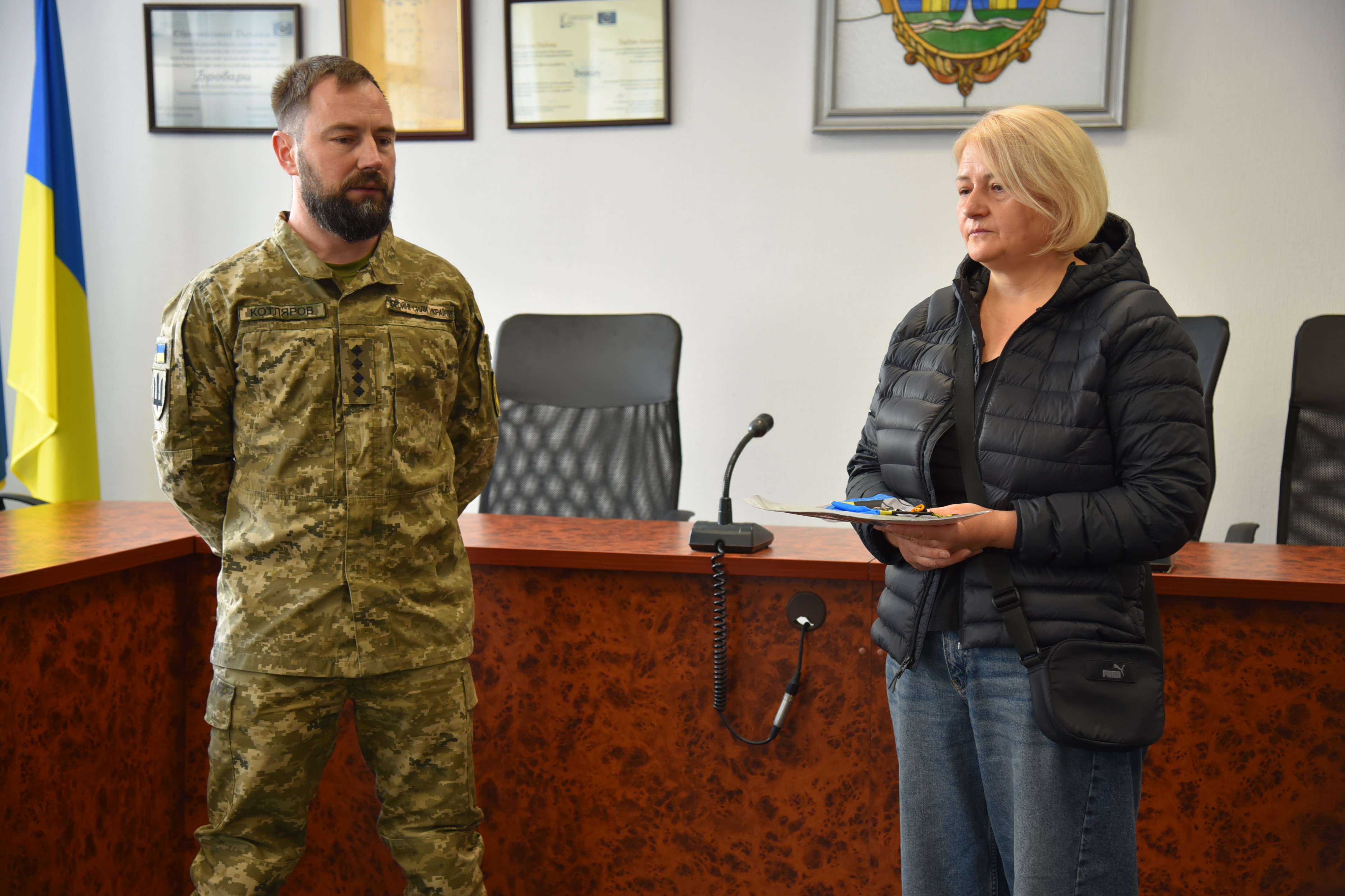 Військовий із руками за спиною стоїть поруч із жінкою, яка тримає документи з синьо-жовтою стрічкою. Ліворуч на задньому плані видно прапор.