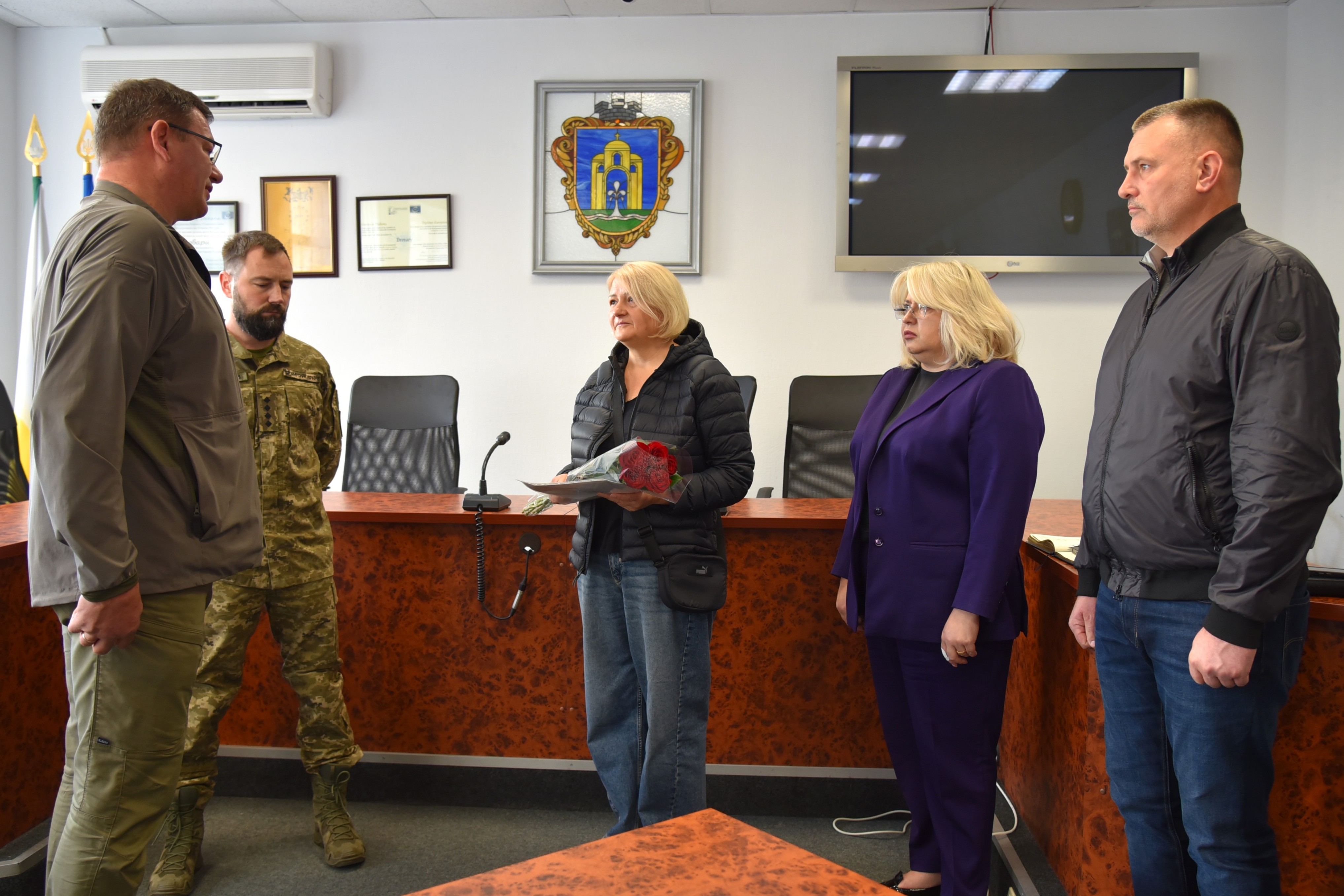 Чоловік у цивільному ліворуч розмовляє з жінкою, яка тримає квіти й документи. Поруч стоять військовий, жінка в фіолетовому костюмі та чоловік.
