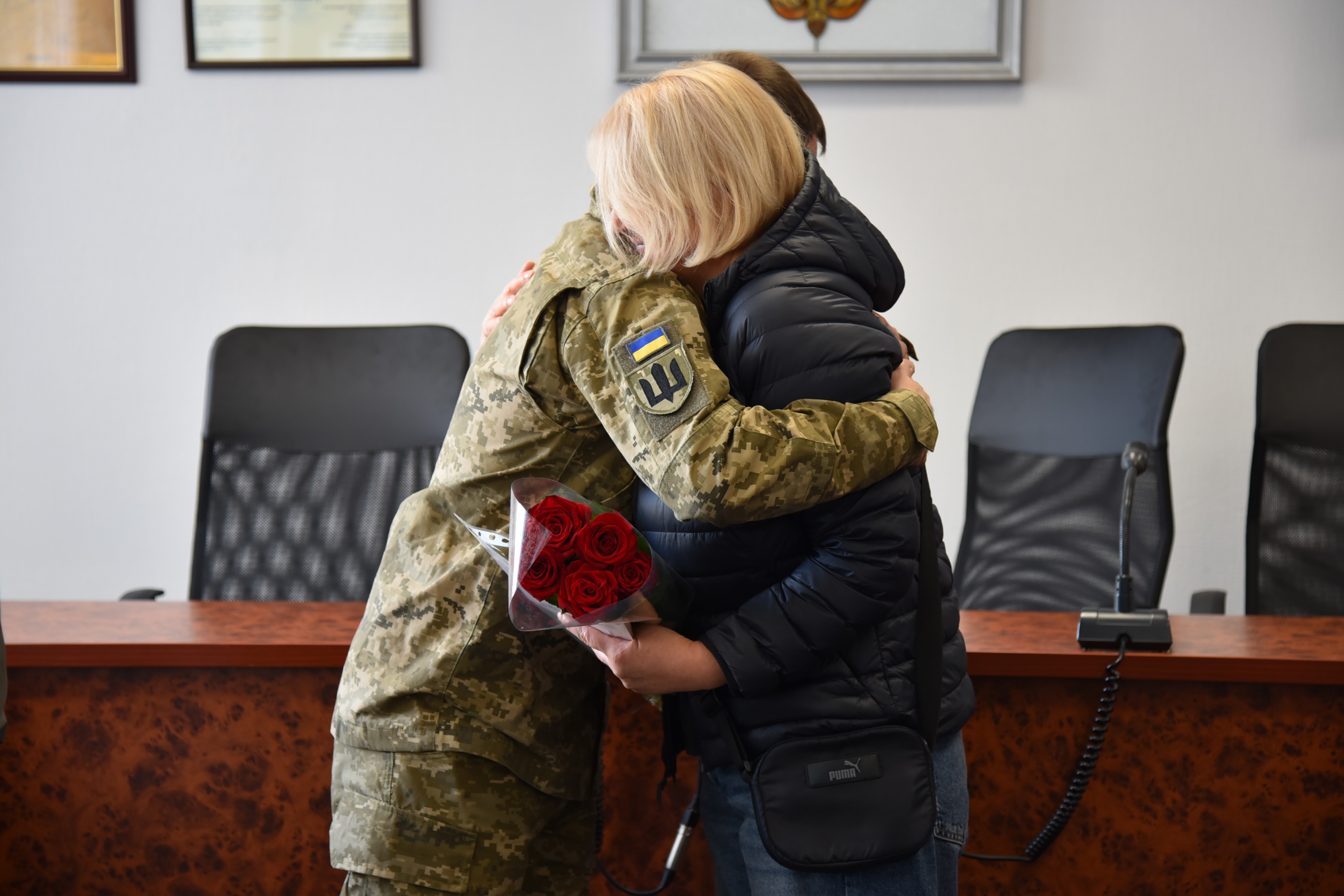 Військовий у камуфляжній формі тепло обіймає жінку в темній куртці, яка тримає букет червоних троянд. На задньому плані офісні стільці.