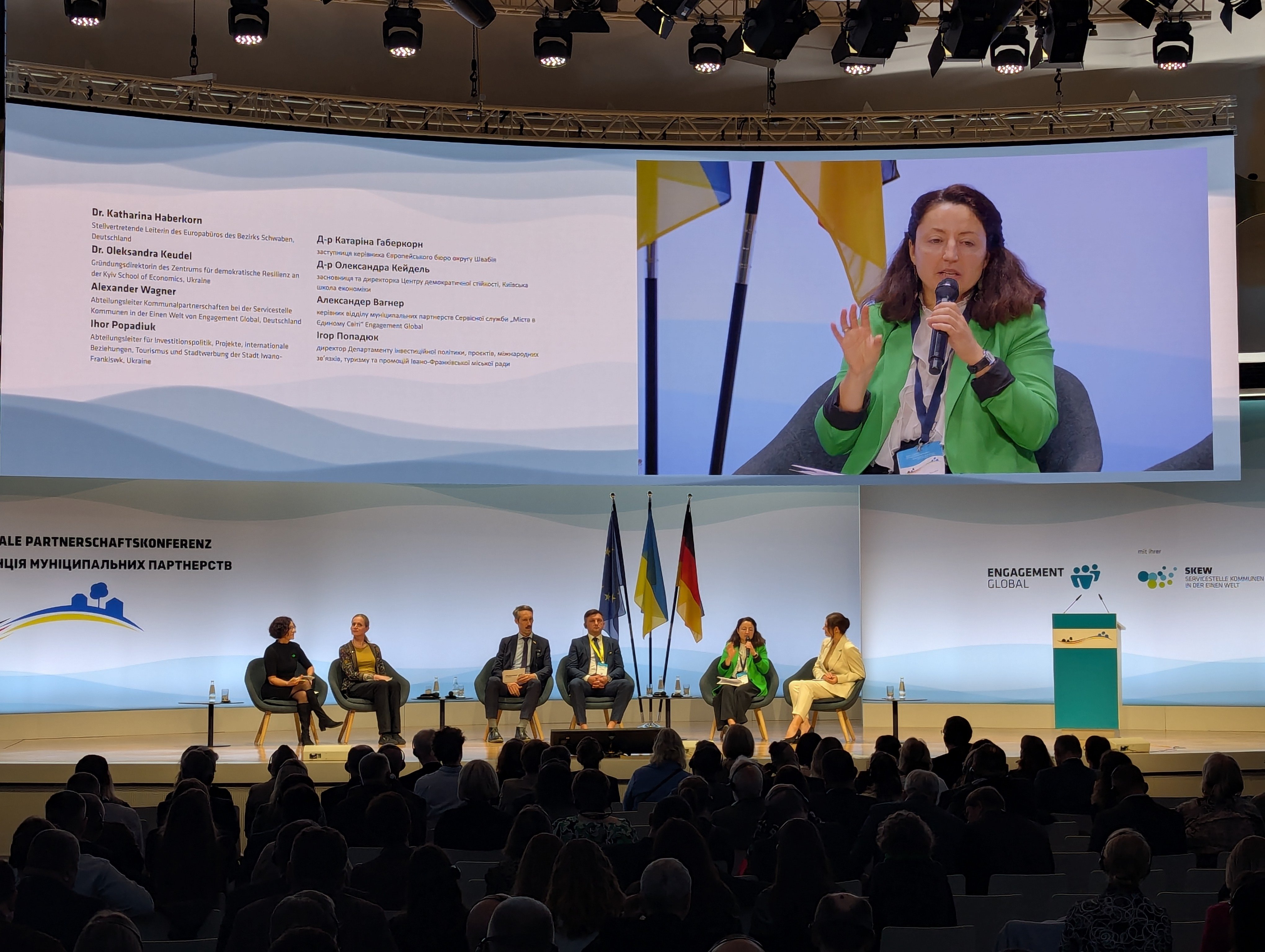 Сцена конференції з великим екраном, де показано спікерку в зеленому піджаці. На сцені сидить панель експертів, попереду — слухачі в залі.