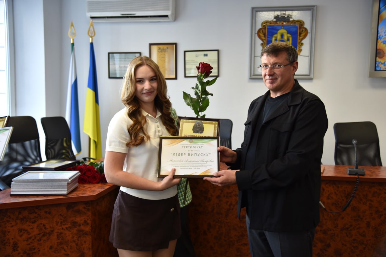Чоловік вільній чорній сорочці вручає дівчині сертифікат «Лідер випуску» та червону троянду в офіційному кабінеті з прапорами України.