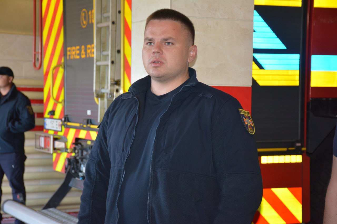Чоловік у формі пожежника стоїть перед пожежною машиною у пожежній частині.
