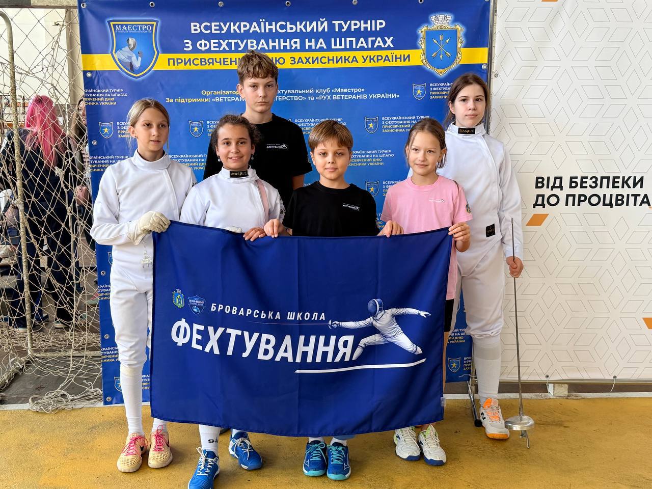 Шість юних спортсменів на тлі банера турніру з фехтування. Попереду діти тримають синій прапор «Броварська школа фехтування».