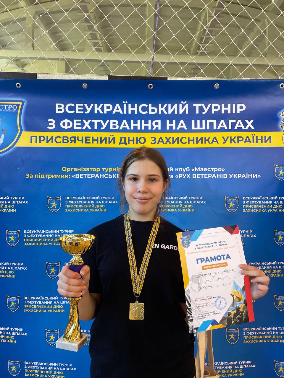 Дівчина-спортсменка з великою золотою медаллю на шиї тримає кубок та грамоту. Вона стоїть на тлі синього банера турніру з фехтування.