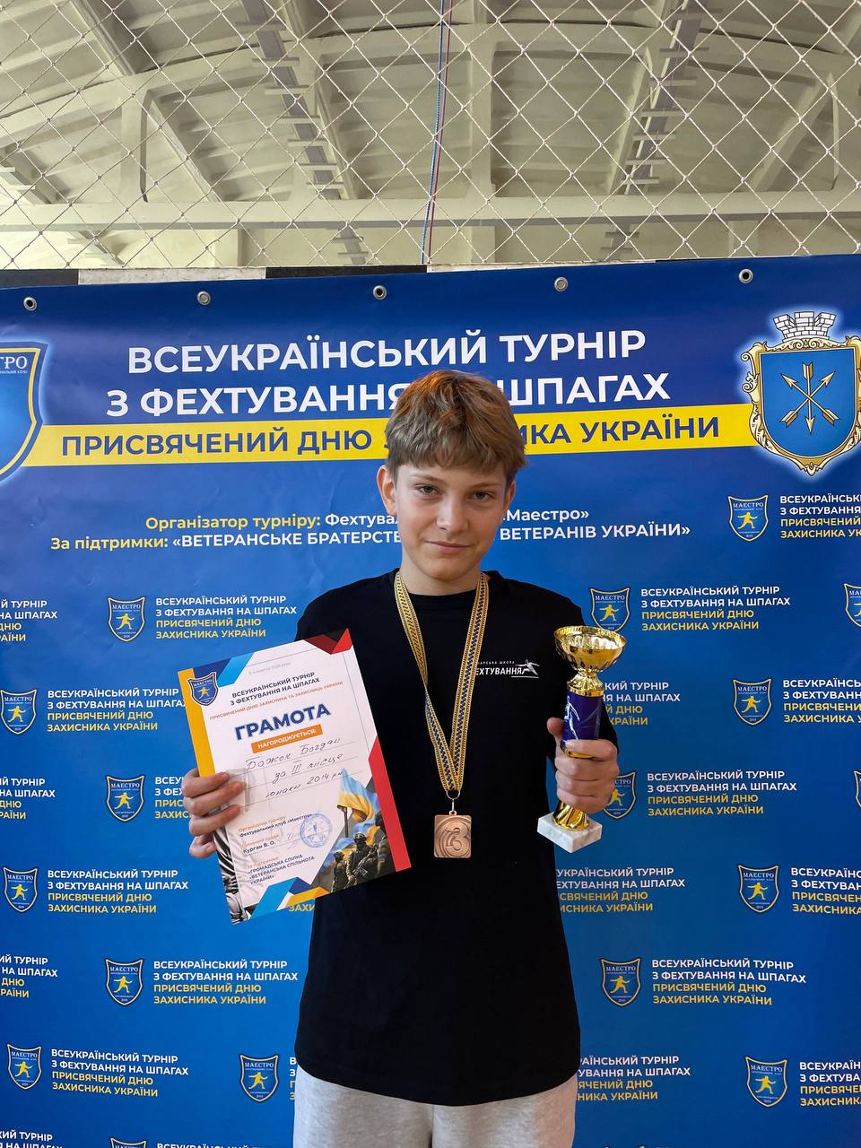 Юний спортсмен із медаллю на шиї стоїть на тлі синього банера турніру. В одній руці він тримає грамоту, а в іншій — золотистий кубок.