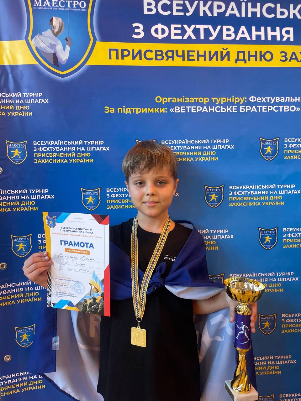 Хлопчик-спортсмен із медаллю на шиї стоїть на тлі синього банера. У руках він тримає кубок та грамоту всеукраїнського турніру з фехтування.