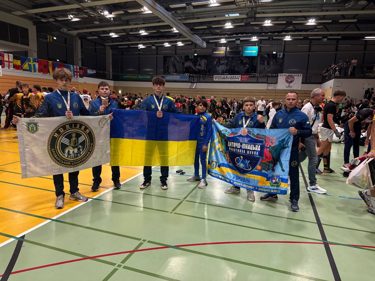 Група юних спортсменів у синій формі стоїть у спортзалі. Вони тримають прапор України та два прапори спортивних клубів Броварів.