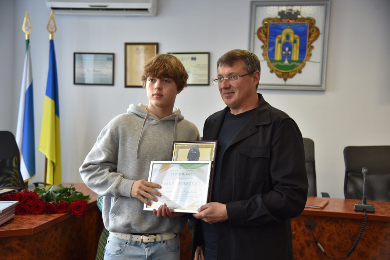 Чоловік у чорній сорочці вручає юнакові в сірому худі сертифікат «Лідер випуску» в офіційному кабінеті з прапорами України.