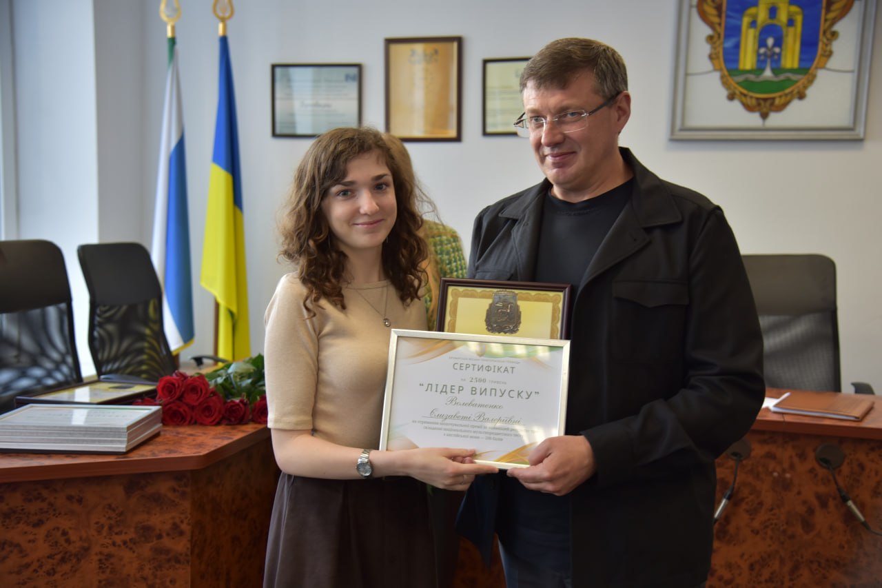 Чоловік у чорній сорочці вручає кучерявій дівчині сертифікат «Лідер випуску» в офіційному кабінеті з прапорами України.