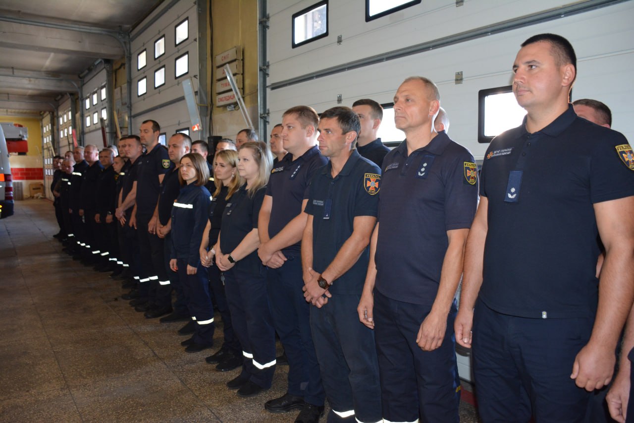 Ряд пожежників у формі стоїть у пожежній частині; позаду видно гаражі та пожежні автомобілі.