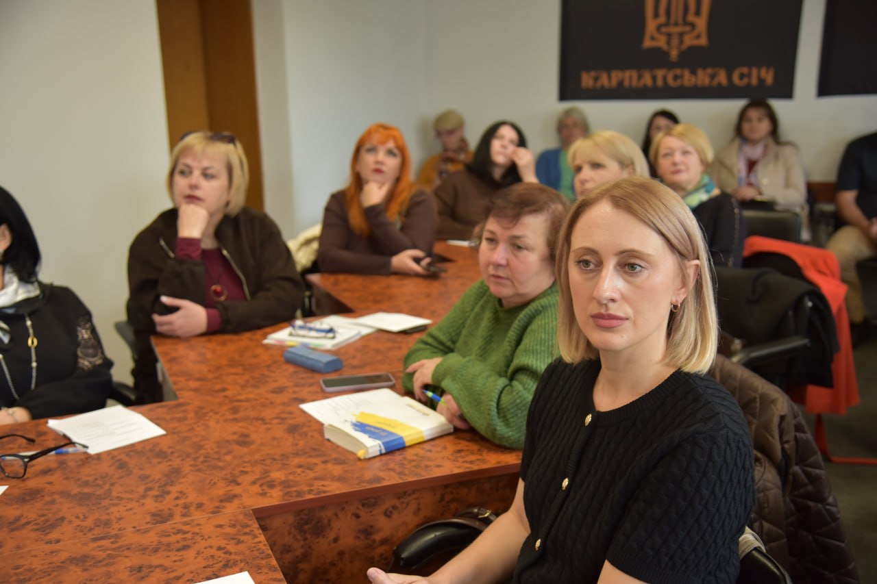Жінка у чорній кофті сидить на передньому плані за столом. Позаду неї інші слухачі та прапор із написом «Карпатська Січ».