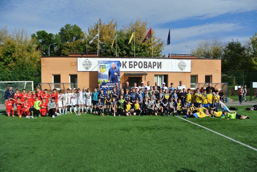 Велика група юних футболістів у різноколірній формі та дорослі стоять на зеленому полі перед будівлею «МФК Бровари» з прапорами на даху.