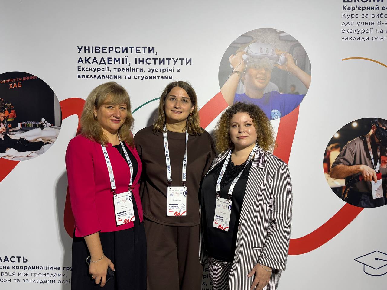 Три жінки з бейджами стоять на тлі банера з написом «УНІВЕРСИТЕТИ, АКАДЕМІЇ, ІНСТИТУТИ» та фотографіями навчального процесу.