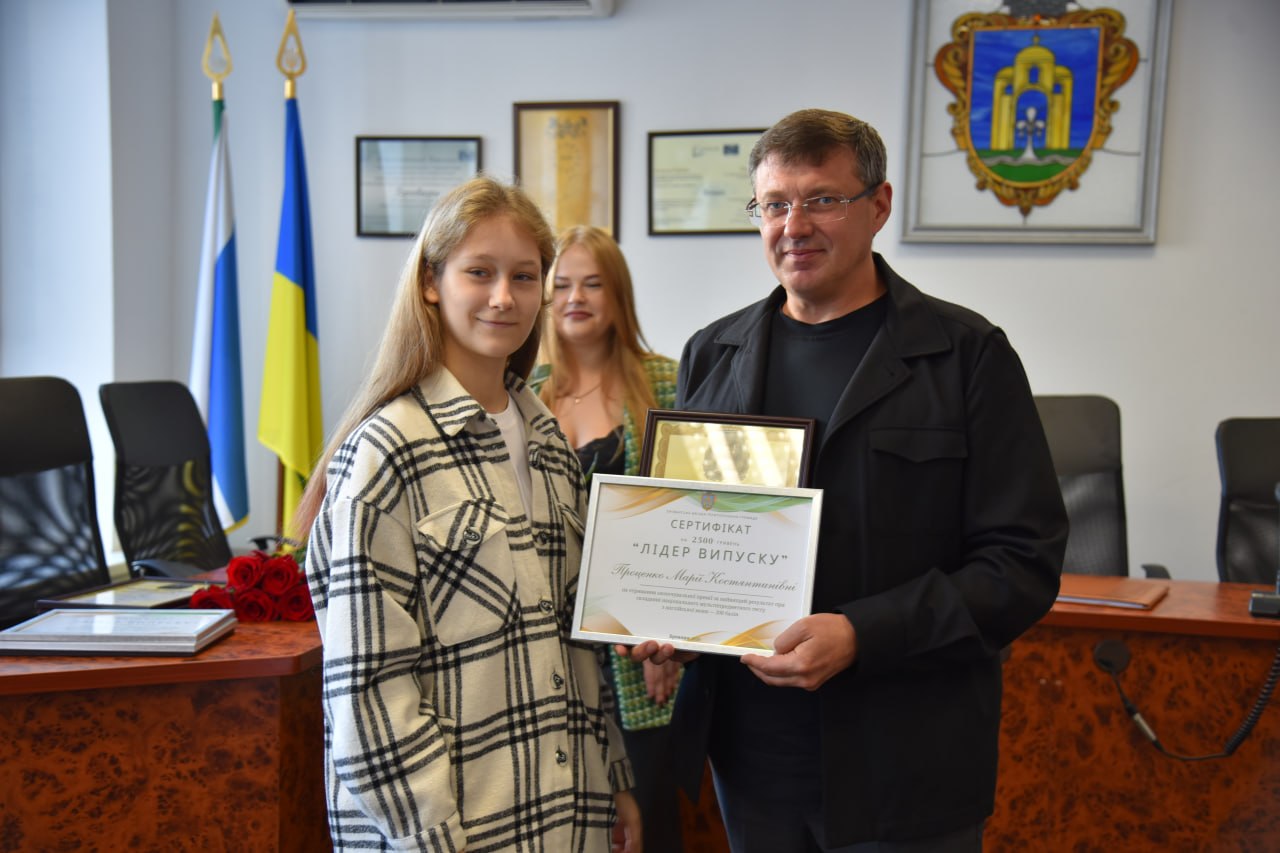 Чоловік у чорній сорочці вручає кучерявій дівчині сертифікат «Лідер випуску» в офіційному кабінеті з прапорами України.