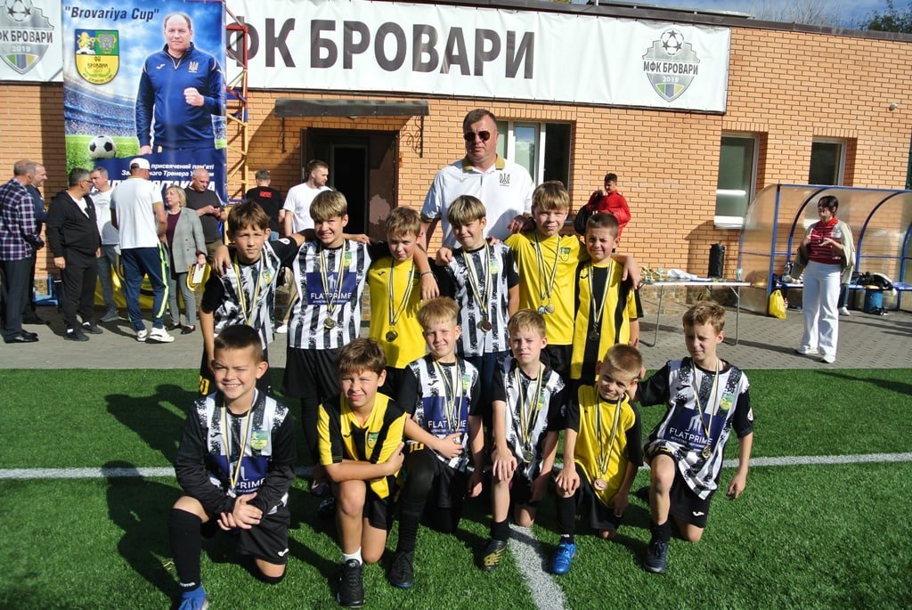 Юні футболісти з медалями на шиї позують на полі. Позаду стоїть тренер, а на тлі будівлі видно напис «ФК БОВАРИ» та банер із фотографією.