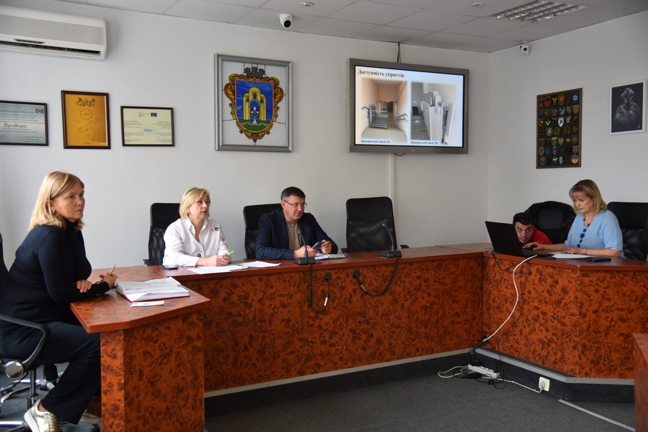 Люди сидять за великим кутовим столом у залі засідань. На стіні висить герб та екран із презентацією «Доступність укриттів».