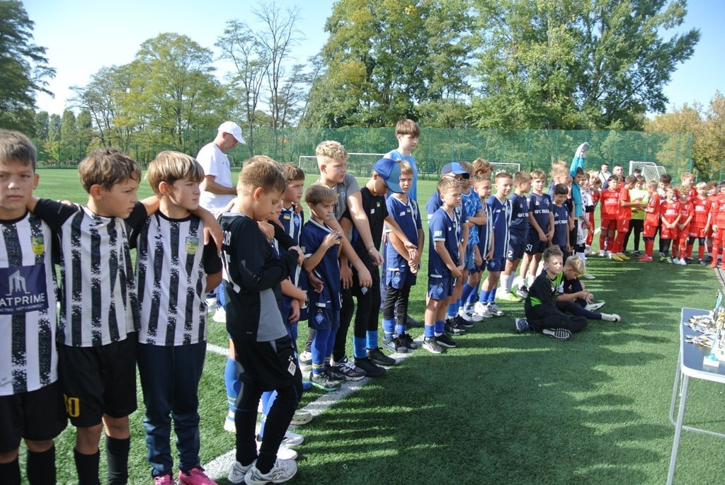 Дві дитячі команди у чорно-білій та синій формі шикуються на полі. Праворуч на столику видно нагороди кубків.