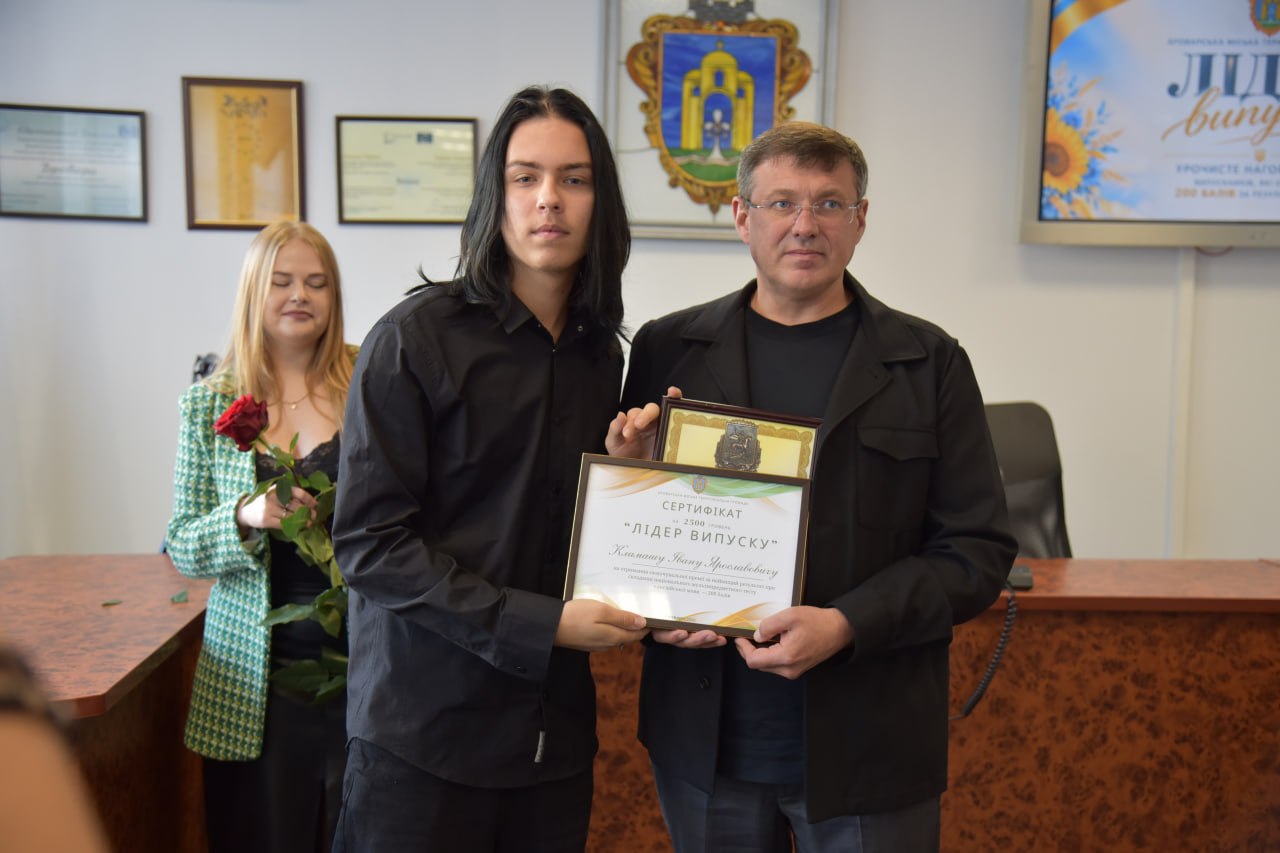 Чоловік у чорній сорочці вручає юнакові в сірому худі сертифікат «Лідер випуску» в офіційному кабінеті з прапорами України.