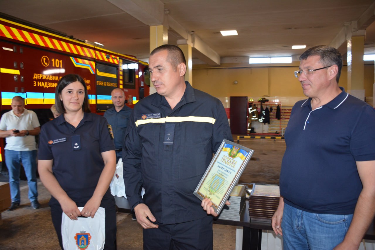 Троє людей у пожежній частині; чоловік у формі тримає подяку, поруч жінка та чоловік, позаду пожежна машина.