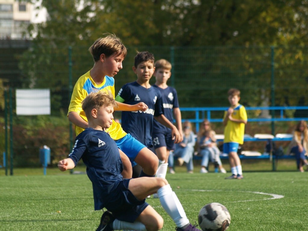 Футбольний матч: гравець у синій формі намагається в підкаті відібрати м'яч у суперника в жовто-блакитній футболці. Позаду стежать інші діти.